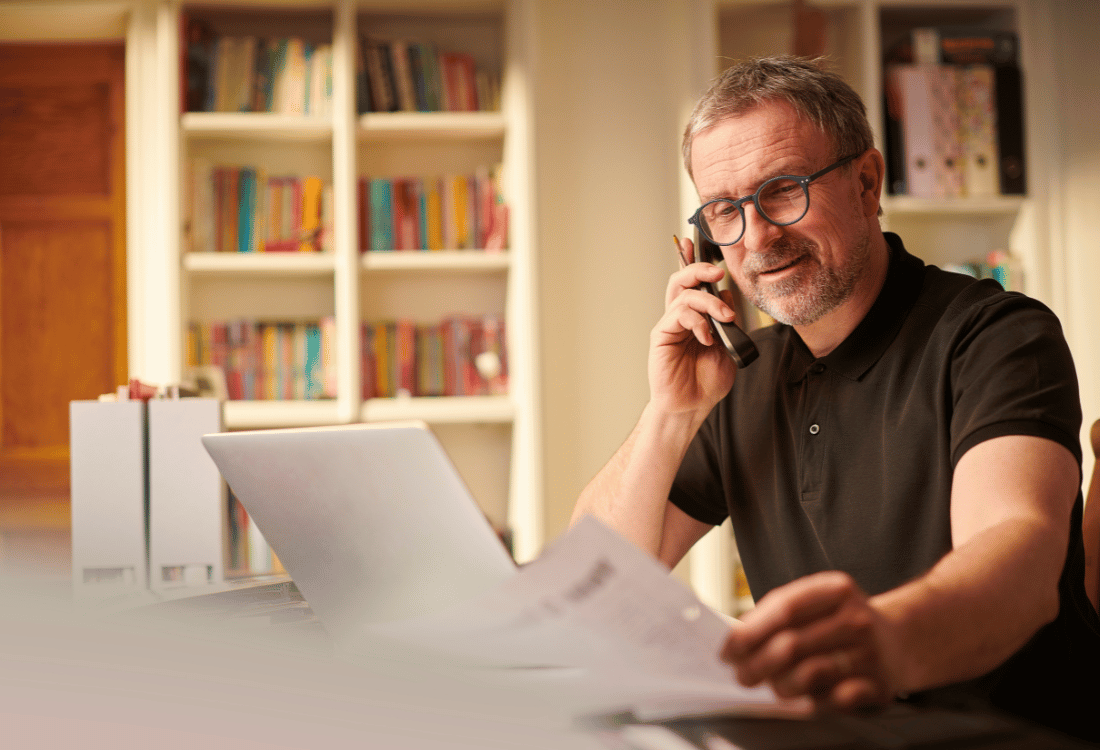 A man in a black shirt working from home, speaking on the phone and holding documents, with a laptop and bookshelf in the background.