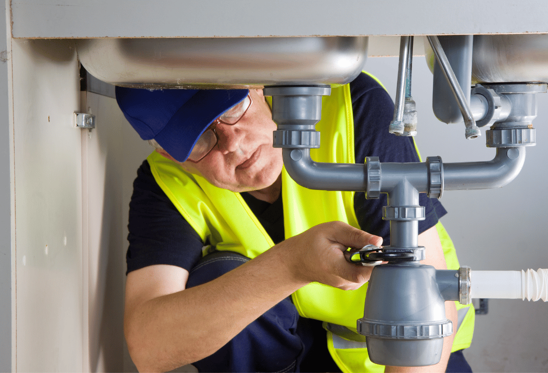 A plumber in a yellow safety vest and blue cap working on pipes under a kitchen sink.