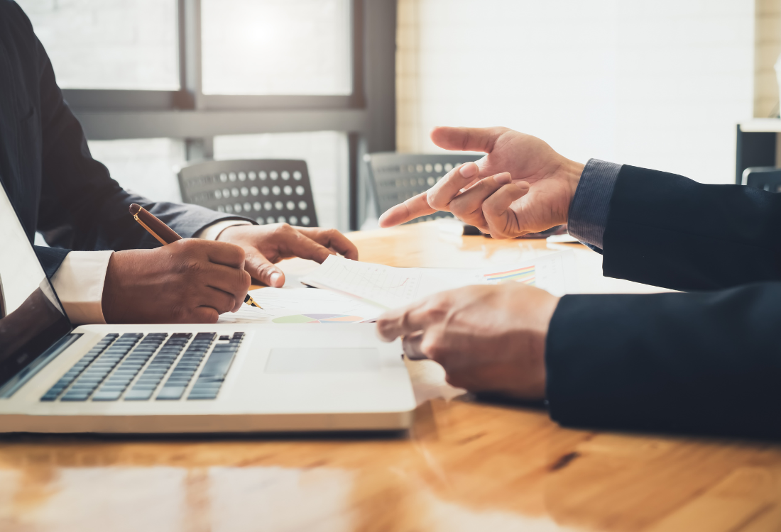 Two people in business attire working together at a desk with a laptop and documents, discussing charts and notes in a modern office setting.